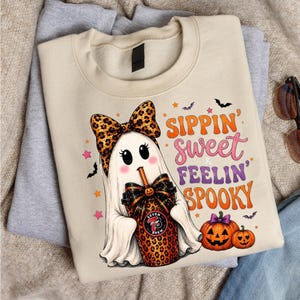 May include: A beige crewneck sweatshirt featuring a graphic of a white ghost wearing a leopard print bow, holding a leopard print cup with a straw. The text reads Sippin' sweet & Feelin' SPOOKY, accompanied by two orange pumpkins and small bats.