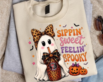 May include: A beige crewneck sweatshirt featuring a graphic of a white ghost wearing a leopard print bow, holding a leopard print cup with a straw. The text reads Sippin' sweet & Feelin' SPOOKY, accompanied by two orange pumpkins and small bats.