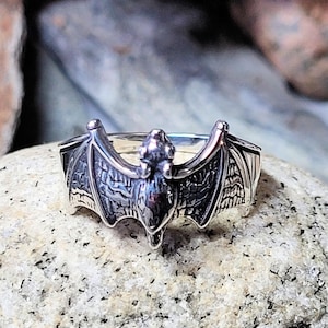 May include: A silver ring with a detailed bat design. The bat's wings are spread out and the ring is set on a gray stone.