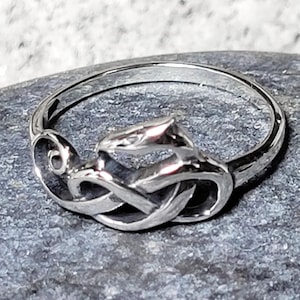 May include: A silver ring featuring a Celtic knot design. The ring has a polished finish and a detailed knot pattern in the center. The ring is set against a gray stone surface.