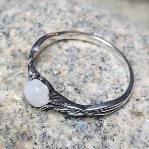 May include: A silver ring with a round, pale gemstone. The ring's band features a detailed, leaf-like design that curves around the stone. The gemstone is a light, opaque color, and the ring is set against a textured background.