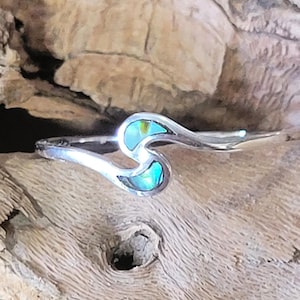 May include: A sterling silver ring with an abstract wave design. The ring features two sections of iridescent abalone shell in shades of blue, green, and yellow. The ring is set against a natural wood background.
