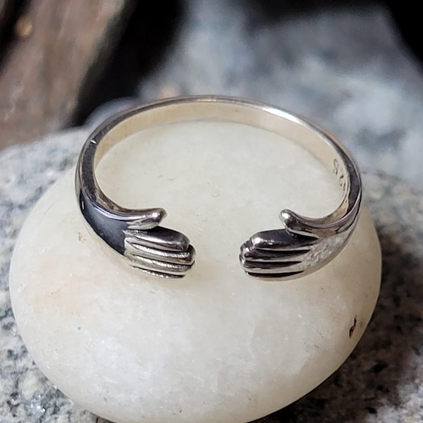 May include: A silver ring with a unique design. The ring features two hands that appear to be embracing. The ring is set on a white stone, with a blurred background.