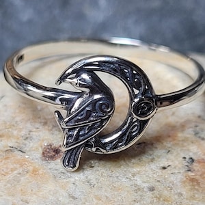 May include: A silver ring featuring a detailed raven perched on a crescent moon. The raven and moon have intricate etched designs. The ring is made of polished metal, with a focus on the bird and moon's symbolic imagery.