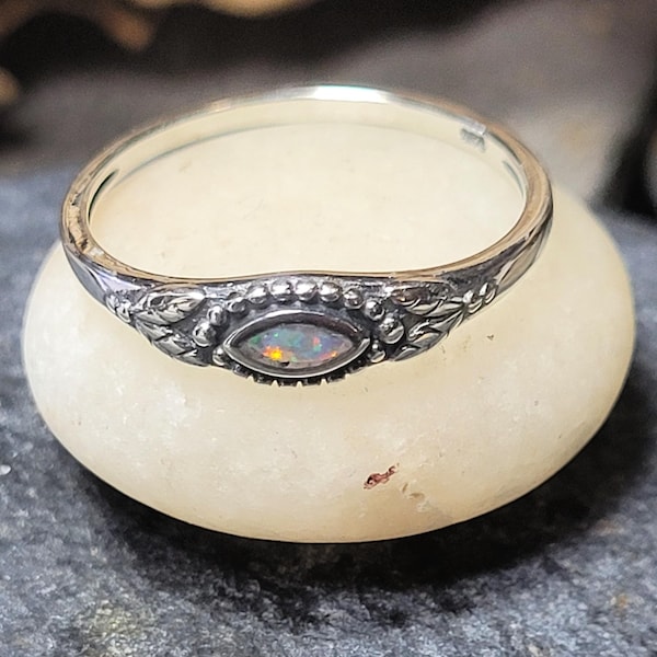 May include: A silver ring with an oval opal gemstone. The ring features a detailed floral design around the gemstone and along the band. The opal displays a play of color, with flashes of green, yellow, and orange. The ring is set on a white stone.