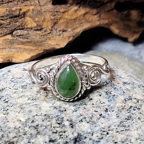 May include: A silver ring with a green jade stone set in a teardrop shape. The ring has a twisted band with a swirl design on each side of the stone.