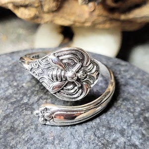 May include: A silver-colored ring with a bee design. The ring features a detailed bee motif on the top and a decorative floral pattern on the band. The ring is adjustable and sits on a gray stone surface.