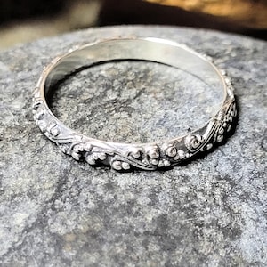 May include: Close-up of a silver ring with an ornate floral design. The ring is highly detailed, featuring swirling patterns and small, raised embellishments. The ring is set against a textured, gray surface.