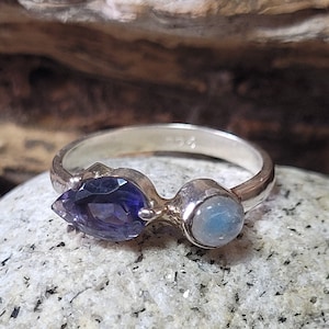 May include: A silver ring with a purple amethyst and a blue moonstone. The ring is set on a gray stone background.