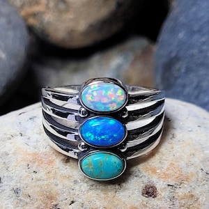 May include: A silver ring featuring three oval gemstones: a top opal with iridescent colors, a vibrant blue stone in the middle, and a turquoise stone at the bottom. The ring has a ribbed band design.