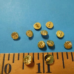 May include: A collection of ten small, gold-colored buttons with a textured surface, scattered on a blue background. A wooden ruler with inch markings is at the bottom of the image.