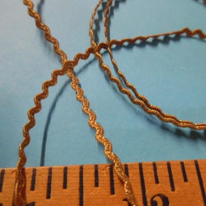 May include: Close-up of gold-colored, wavy trim ribbon. The ribbon is lying on a blue surface next to a wooden ruler. The ribbon has a zig-zag pattern and appears to be made of a shiny material. The ruler is marked in inches.