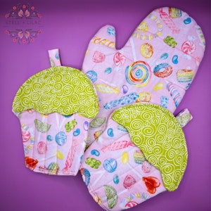 May include: Set of three pink and green oven mitts with a candy theme. The mitts are shaped like cupcakes and feature a variety of colorful candies, lollipops, and sprinkles.