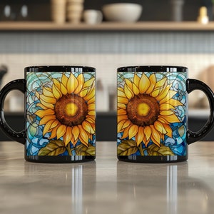 May include: Two black ceramic mugs featuring a vibrant sunflower design with yellow petals and a brown center, set against a blue stained-glass style background. Each mug has a curved black handle and a smooth, glossy finish on the exterior.
