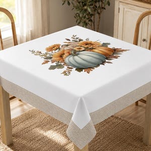 May include: A square white tablecloth featuring a central illustration of a light blue pumpkin surrounded by orange sunflowers, autumn leaves, and smaller pumpkins. The tablecloth has a tan and white checkered border along the bottom edge.