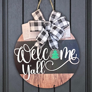 May include: A round wooden door hanger with a black and white checkered bow. The sign says "Welcome Y'all" in white lettering with a green Christmas tree cutout.