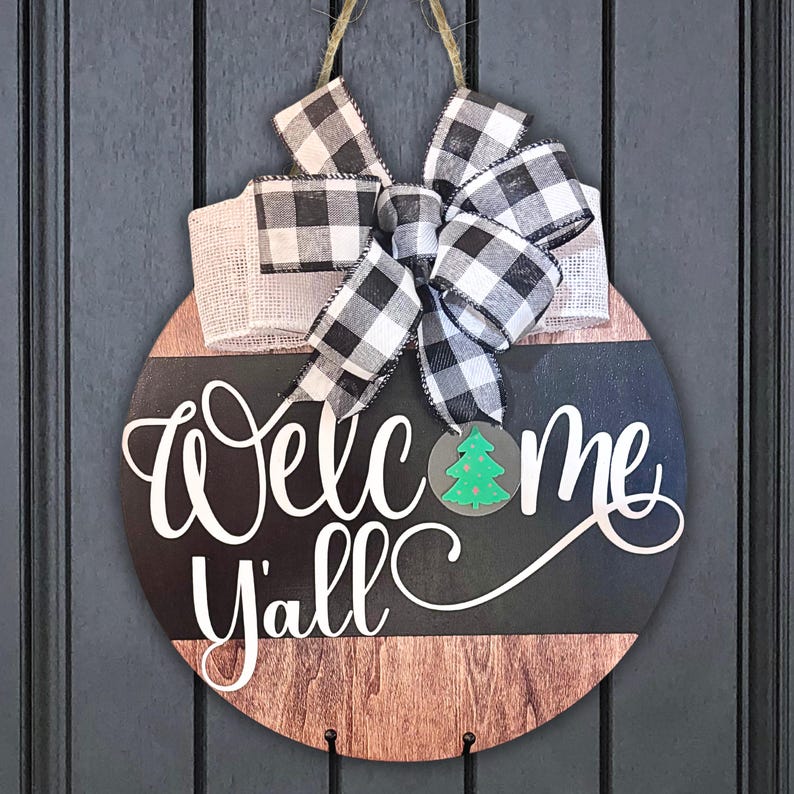 May include: A round wooden door hanger with a black and white checkered bow. The sign says "Welcome Y'all" in white lettering with a green Christmas tree cutout.