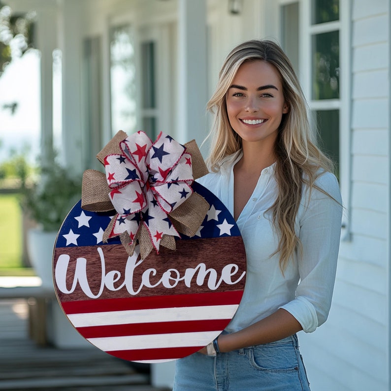 May include: A round wooden welcome sign with a patriotic theme. The sign features a blue section with white stars, red and white stripes, and the word "Welcome" in white script. A decorative bow with red, white, and blue stars and burlap accents tops the sign.