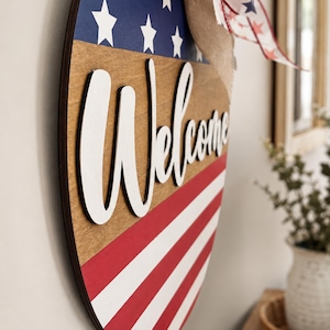 K&ouml;nnte beinhalten: Willkommensschild aus Holz mit patriotischem Thema. Das schildf&ouml;rmige Schild zeigt rot-wei&szlig;e Streifen, einen blauen Bereich mit wei&szlig;en Sternen und das Wort "Welcome" in wei&szlig;er Schrift.