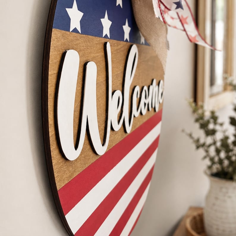 K&ouml;nnte beinhalten: Willkommensschild aus Holz mit patriotischem Thema. Das schildf&ouml;rmige Schild zeigt rot-wei&szlig;e Streifen, einen blauen Bereich mit wei&szlig;en Sternen und das Wort "Welcome" in wei&szlig;er Schrift.