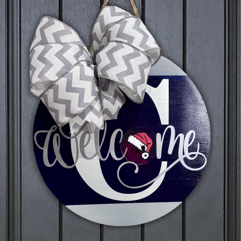 May include: A round wooden door hanger with a white letter "C" and the word "welcome" in cursive script. The letter "C" has a red Santa hat on top. The door hanger is decorated with a gray and white chevron ribbon.