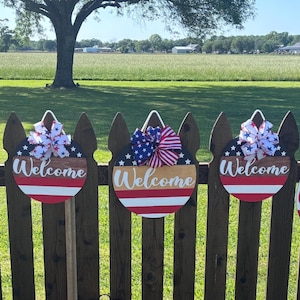 K&ouml;nnte beinhalten: Eine Reihe patriotischer Willkommensschilder in Rot, Wei&szlig; und Blau. Jedes Schild hat einen Sternenhimmel, eine dekorative Schleife und den Schriftzug "Welcome". Ein Schild tr&auml;gt den Namen "Wilcox".