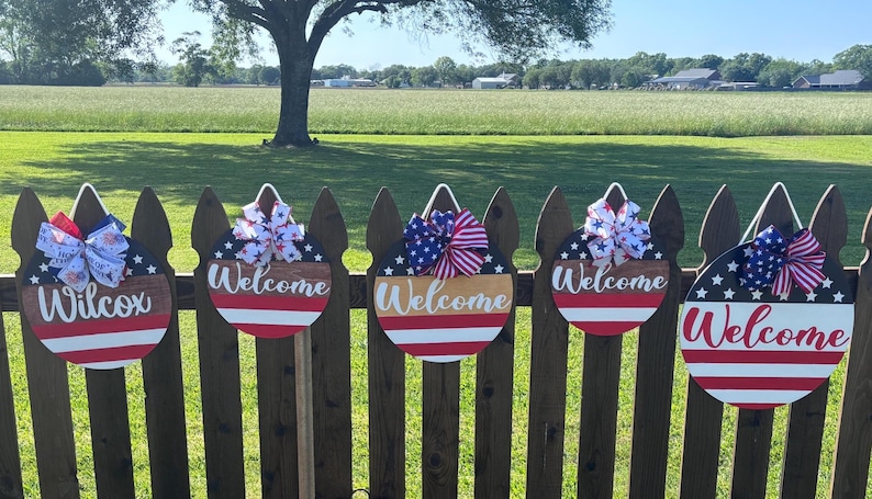 K&ouml;nnte beinhalten: Eine Reihe patriotischer Willkommensschilder in Rot, Wei&szlig; und Blau. Jedes Schild hat einen Sternenhimmel, eine dekorative Schleife und den Schriftzug "Welcome". Ein Schild tr&auml;gt den Namen "Wilcox".