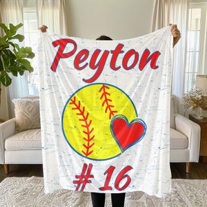 May include: White blanket with a softball design. The blanket features the name "Peyton" in red script, a yellow softball with red stitching, a red heart, and the number "16" in red.