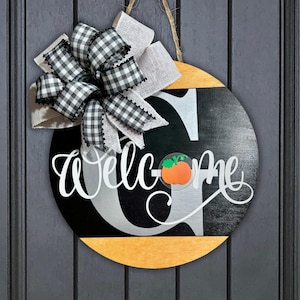 May include: A black and white welcome sign with a silver letter G and a small orange pumpkin. The sign is decorated with a black and white checkered bow and a burlap bow.