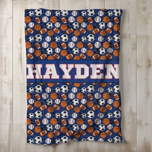 Könnte beinhalten: Eine blaue Decke mit einem Muster aus Sportbällen, darunter Fußbälle, Basketbälle und Basebälle. Der Name "HAYDEN" ist in weißen Blockbuchstaben auf einem blauen Streifen abgebildet. Die Decke ist aus weichem Material.