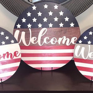 K&ouml;nnte beinhalten: Drei runde Holzschilder mit patriotischem Thema. Jedes Schild zeigt ein Design, das von der amerikanischen Flagge inspiriert ist, mit Sternen, Streifen und dem Wort "Welcome" in einer Schrift. Die Schilder werden auf einer dunklen Oberfl&auml;che pr&auml;sentiert.