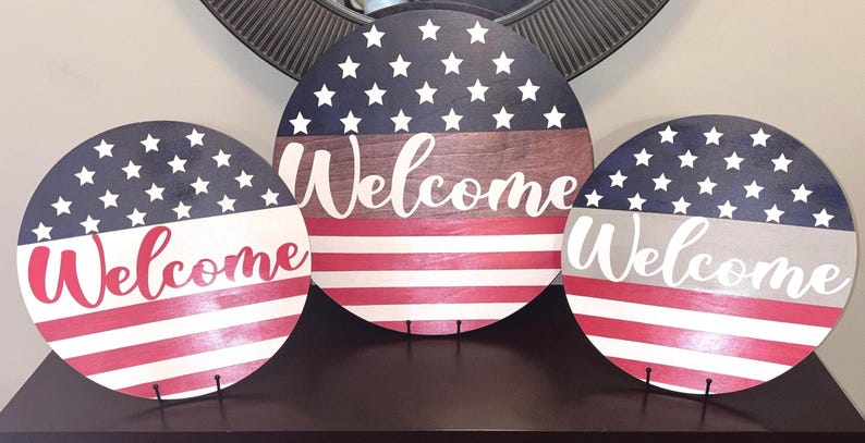 K&ouml;nnte beinhalten: Drei runde Holzschilder mit patriotischem Thema. Jedes Schild zeigt ein Design, das von der amerikanischen Flagge inspiriert ist, mit Sternen, Streifen und dem Wort "Welcome" in einer Schrift. Die Schilder werden auf einer dunklen Oberfl&auml;che pr&auml;sentiert.