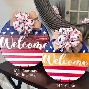 K&ouml;nnte beinhalten: Zwei runde Holzschilder mit patriotischem Thema. Eines hat einen Durchmesser von 45,7 cm, das andere von 35,6 cm. Beide zeigen eine "Welcome"-Nachricht, sternenbesetzte Schleifen und rot, wei&szlig; und blaue Farben.