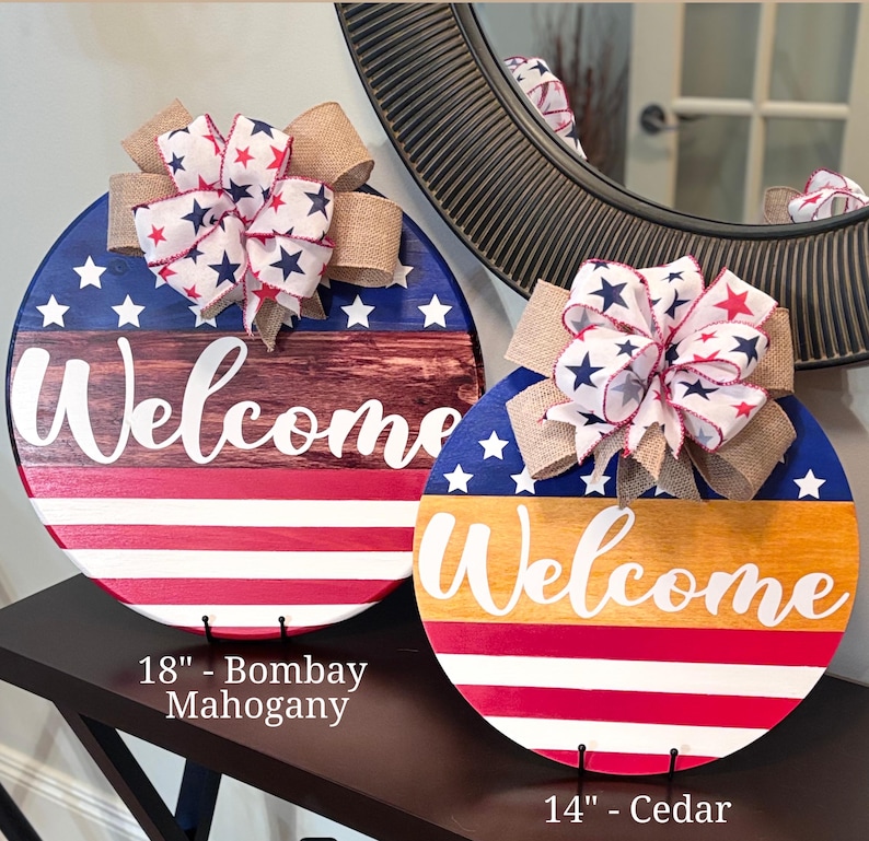 K&ouml;nnte beinhalten: Zwei runde Holzschilder mit patriotischem Thema. Eines hat einen Durchmesser von 45,7 cm, das andere von 35,6 cm. Beide zeigen eine "Welcome"-Nachricht, sternenbesetzte Schleifen und rot, wei&szlig; und blaue Farben.