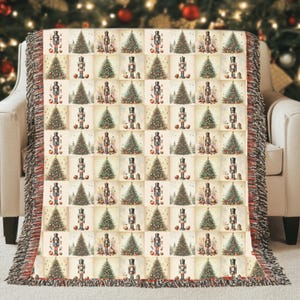 May include: A cream-colored throw blanket with a repeating pattern of Christmas trees and nutcrackers. The blanket has a red and black fringed edge and is draped over a beige armchair. The background is a blurred Christmas tree.