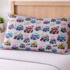 May include: A rectangular pillowcase with a gray background, featuring colorful monster truck illustrations and the name "Liam" in a playful font. The trucks are in various colors, with splashes of paint.