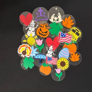 May include: A collection of 20 clear plastic circles with various colorful holiday and seasonal images. The images include a snowman, a pumpkin, a heart, a shamrock, a turkey, a sunflower, a school bus, a cross, a star, a tulip, an umbrella, an egg, a leaf, and an American flag.