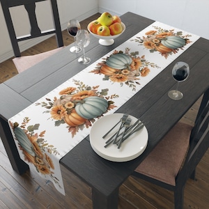 Puede incluir: Un camino de mesa blanco con un tema otoñal que presenta un patrón repetido de calabazas naranjas y azules con girasoles y hojas. El camino de mesa está colocado sobre una mesa de madera oscura con un tazón de manzanas, copas de vino y una mesa puesta.
