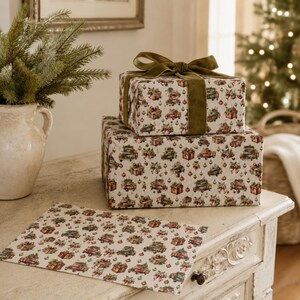 May include: Two rectangular gift boxes stacked and wrapped in cream-colored paper featuring a repeating pattern of vintage red and green trucks carrying Christmas trees and presents, tied with a wide olive green velvet ribbon and bow.