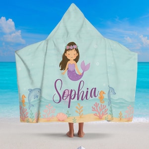 May include: A hooded beach towel featuring a mermaid illustration with the name "Sophia" in purple script. The towel is light blue with a sea-themed design, including dolphins, seahorses, and coral. The towel is designed to be worn.