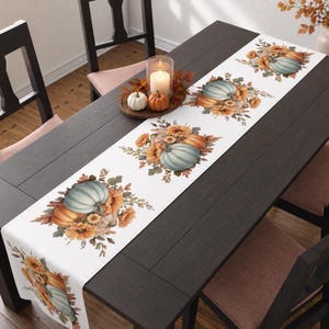 May include: A white table runner featuring a repeating pattern of light blue and orange pumpkins, yellow sunflowers, and autumn leaves. The runner is laid out on a dark wooden table, with a wooden tray holding two small pumpkins and a candle.