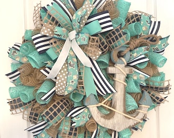May include: A nautical-themed wreath featuring a wooden anchor and various ribbons. The wreath incorporates teal, navy, and tan mesh and ribbons with star and seashell patterns. The anchor is painted in shades of blue and white, with rope detailing.