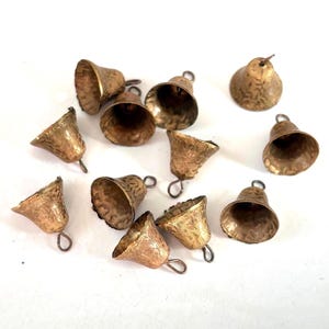 Puede incluir: Un conjunto de once pequeñas campanas de metal en tono dorado. Cada campana presenta una superficie texturizada con un patrón grabado y un pequeño lazo en la parte superior para colgar. Las campanas están dispuestas en formación dispersa.