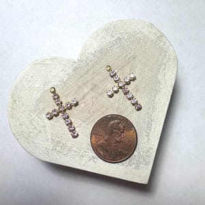 Puede incluir: Dos colgantes dorados en forma de cruz adornados con pedrería rosa descansan sobre una superficie en forma de corazón de color gris claro. Una moneda está al lado de las cruces para comparar el tamaño. Las cruces tienen un pequeño lazo en la parte superior.