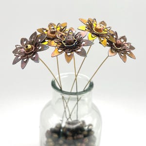 May include: A set of five metal flowers with layered, cutout petals in shades of bronze, copper, and gold. Each flower features a central bead and is attached to a thin wire stem. The flowers are arranged inside a clear glass bottle.