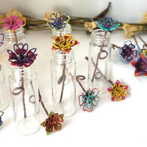 Puede incluir: Una colecci&oacute;n de peque&ntilde;as botellas de vidrio transparente, cada una coronada con una flor colorida con marco de alambre. Las flores son de varios colores, incluyendo rosa, azul, amarillo y morado. Los tallos son marrones y las flores est&aacute;n dispuestas sobre una superficie blanca.