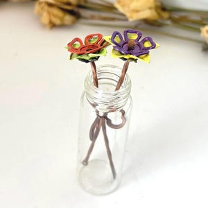 Puede incluir: Una botella de vidrio transparente contiene dos horquillas para el pelo con forma de flor hechas a mano. Una flor es roja y naranja, la otra es morada y amarilla. Los tallos son marrones y las flores tienen hojas verdes. La botella est&aacute; sobre una superficie blanca.