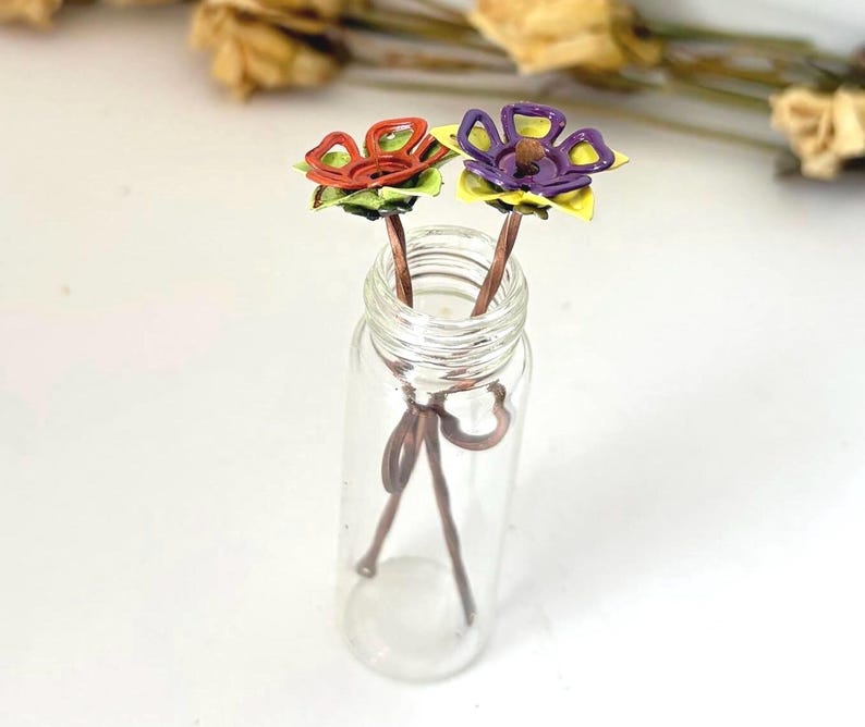 Puede incluir: Una botella de vidrio transparente contiene dos horquillas para el pelo con forma de flor hechas a mano. Una flor es roja y naranja, la otra es morada y amarilla. Los tallos son marrones y las flores tienen hojas verdes. La botella est&aacute; sobre una superficie blanca.