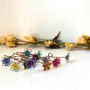 Puede incluir: Una colecci&oacute;n de adornos florales coloridos envueltos en alambre en varios colores, incluyendo azul, amarillo y rosa. Las flores est&aacute;n dispuestas sobre finos tallos marrones. Flores amarillas secas en el fondo.