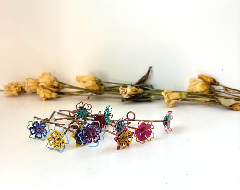 Puede incluir: Una colecci&oacute;n de adornos florales coloridos envueltos en alambre en varios colores, incluyendo azul, amarillo y rosa. Las flores est&aacute;n dispuestas sobre finos tallos marrones. Flores amarillas secas en el fondo.